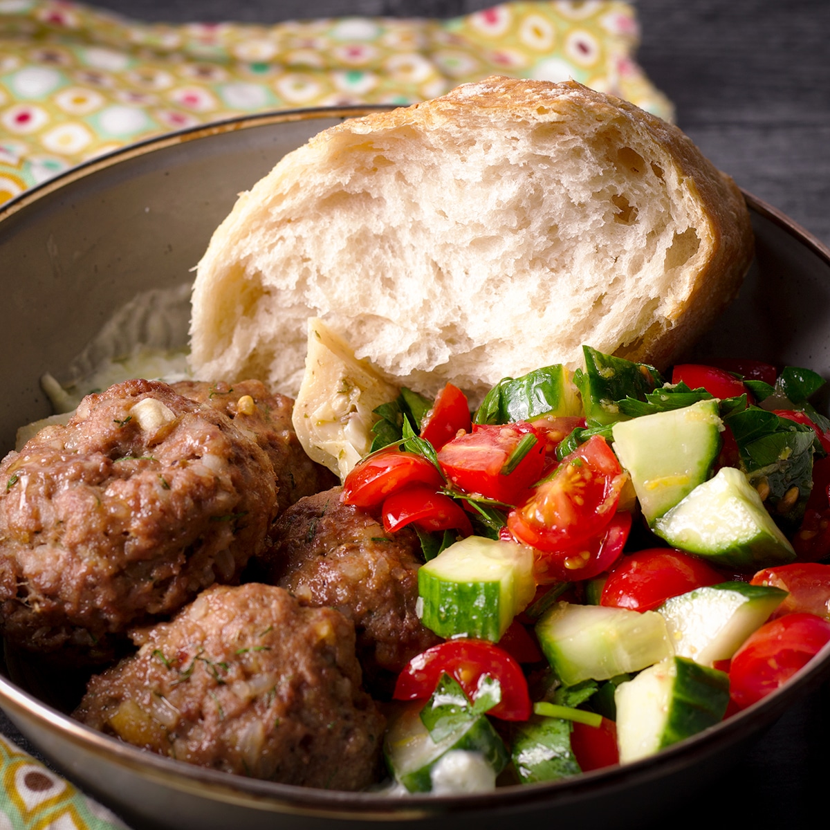 A bowl containing Greek meatballs and tzatziki sauce, and tomato and cucumber salad, and a hunk of white bread.