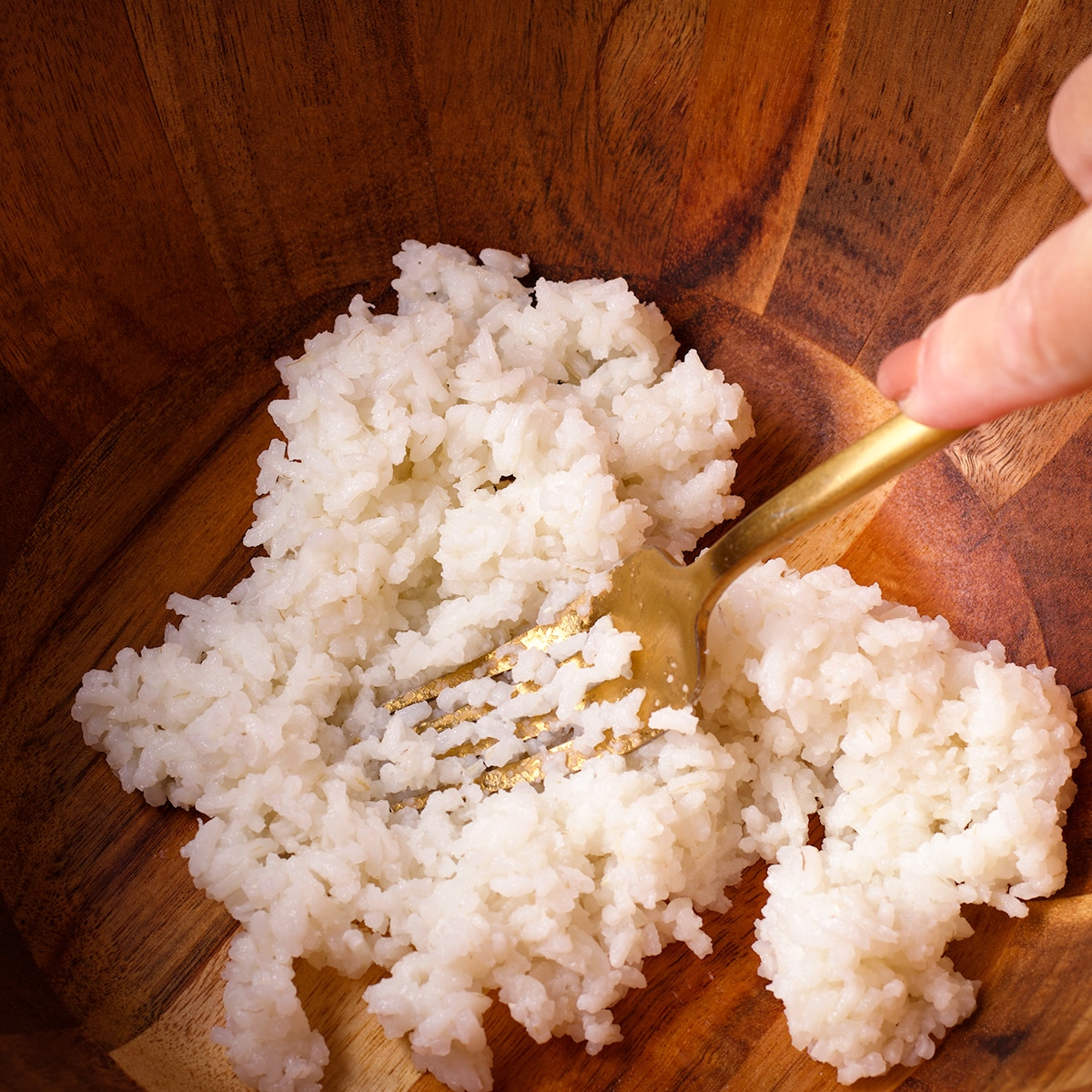 Someone using a fork to mash cooked white rice in a wood bowl.