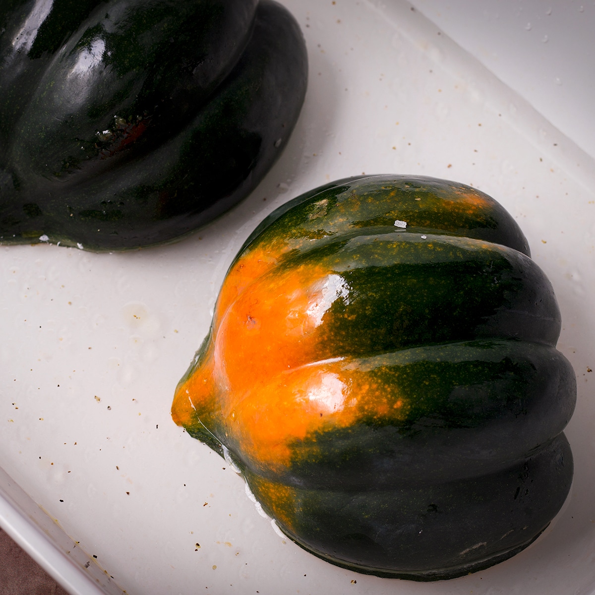 An acorn squash that's been cut in half and placed cut side down in a white baking dish filled with about ½-inch of water.