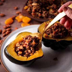 Someone using a gold spoon to fill the center of a roasted acorn squash with cooked quinoa, roasted nuts, and dried fruit.