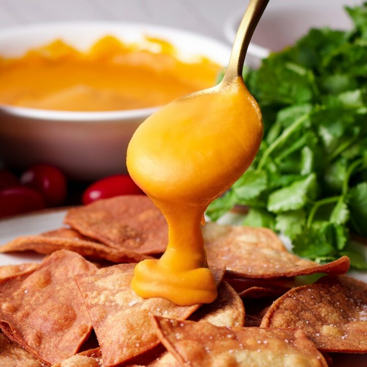 Someone using a gold spoon to pour vegan nacho cheese sauce over a plate full of tortilla chips.