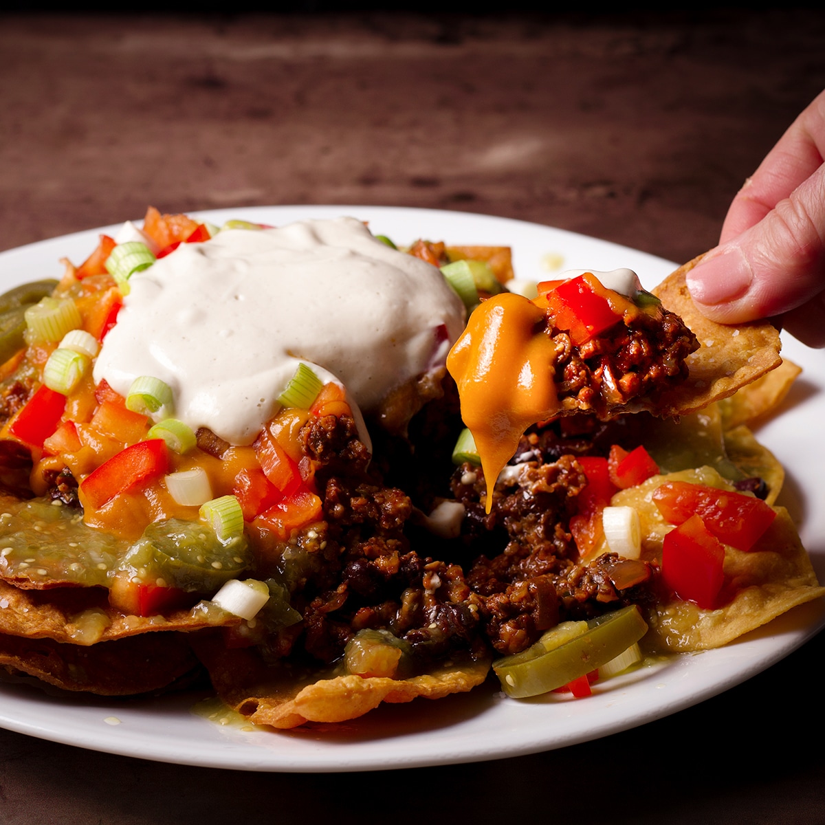 Someone lifting a tortilla chip loaded with vegan taco meat and vegan nacho cheese sauce from a plate of vegan nachos.