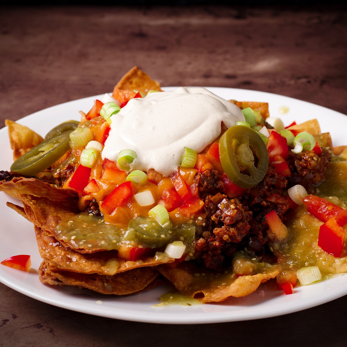 A plate of vegan nachos made from homemade tortilla chips, vegan taco meat, vegan sour cream, vegan cheese sauce, and chopped vegetables.
