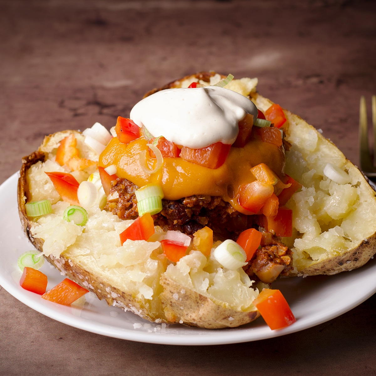 A baked potato split in half and topped with vegan taco meat, vegan nacho cheese, vegan sour cream, and chopped veggies.