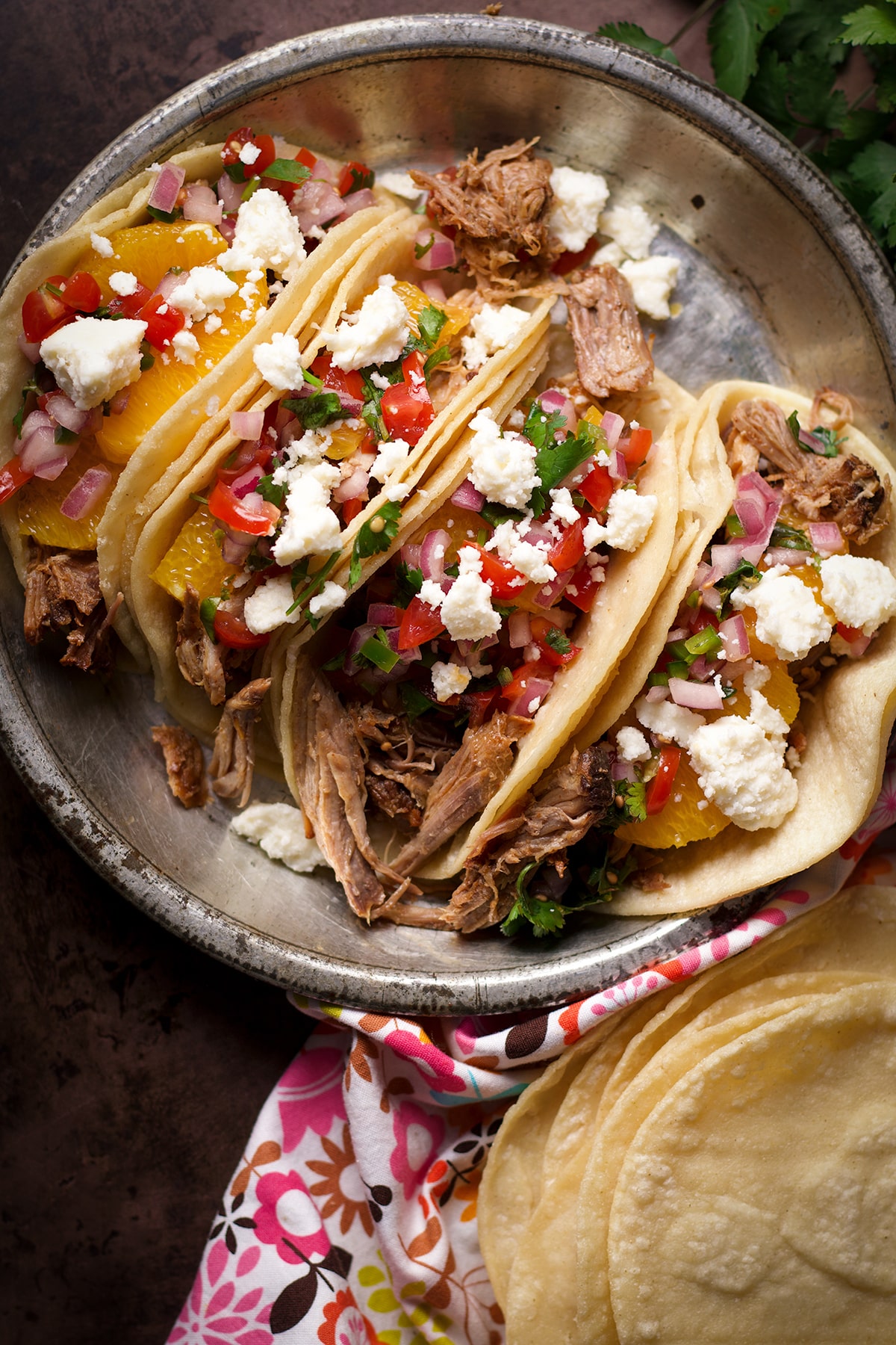 Four shredded pork tacos with orange slices, pico de gallo, and crumbled queso fresco on a tin plate.