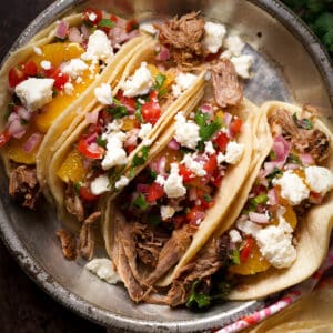 Four shredded pork tacos with orange slices, pico de gallo, and crumbled queso fresco on a tin plate.