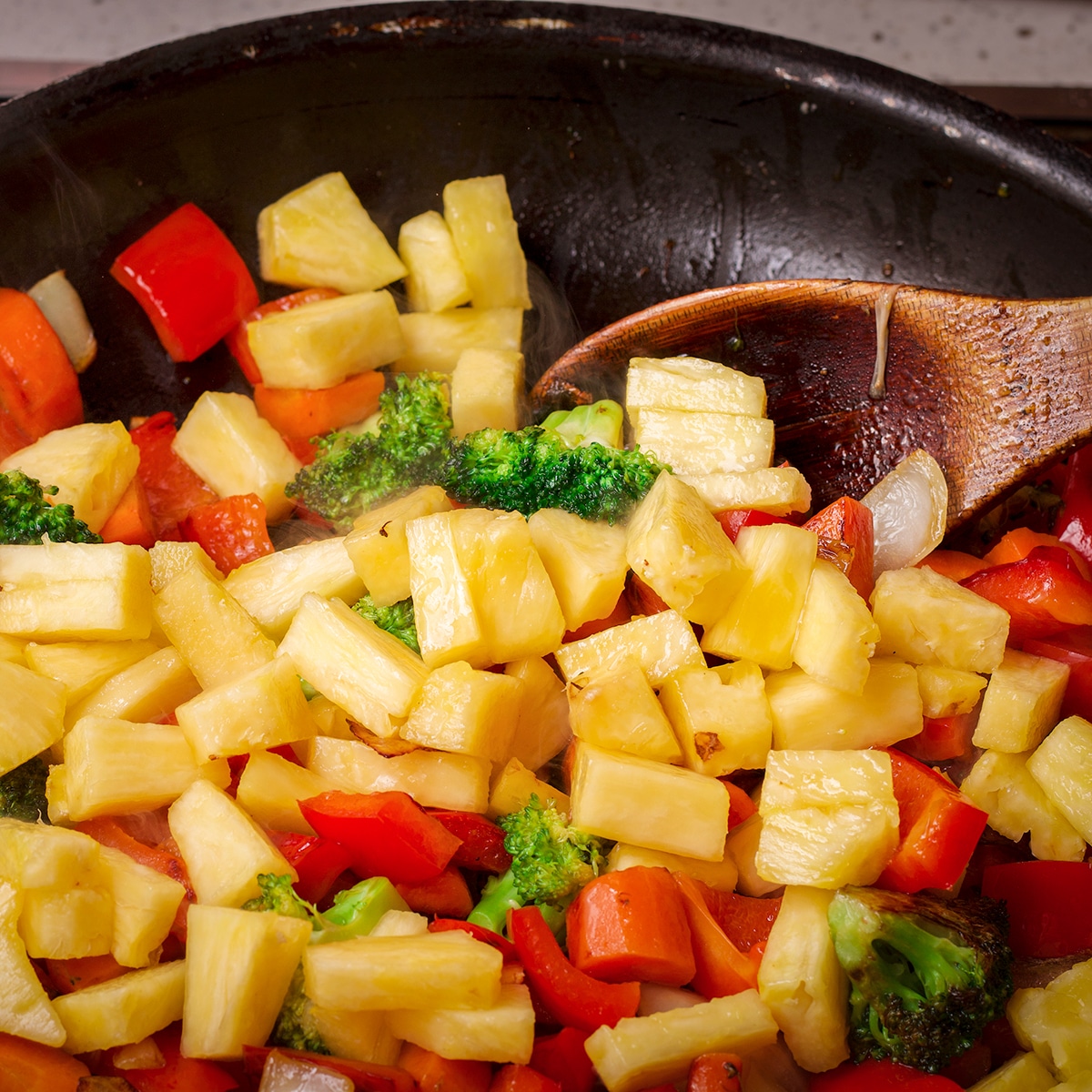 Using a wooden spoon to stir fresh pineapple into vegetables as they cook in a skillet.