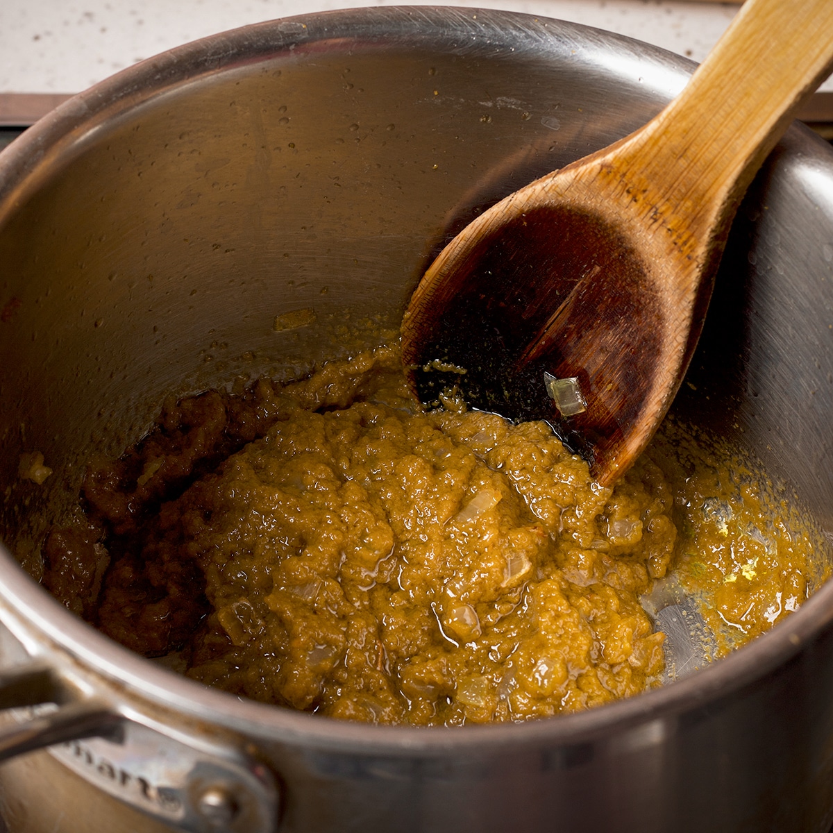 Using a wooden spoon to stir green curry paste into cooked onions and shallots in a saucepan.