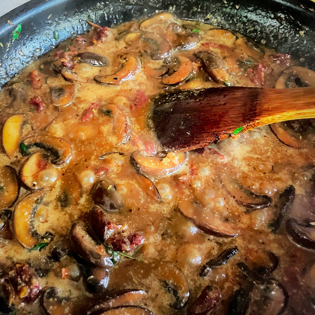 Using a wooden spoon to stir coconut milk and vegetable broth into mushrooms and sun-dried tomatoes as they cook in a hot skillet. 