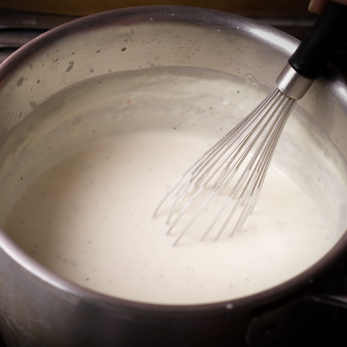 Someone using a wire whisk to stir béchamel sauce while it simmers in a saucepan.