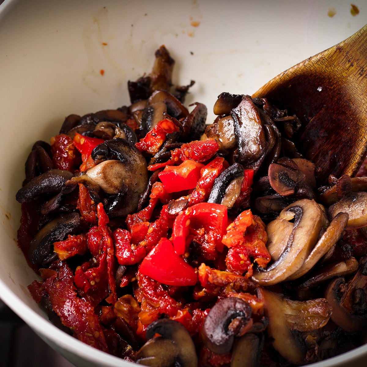 A white bowl containing cooked mushrooms, chopped roasted red peppers, and sun dried tomatoes.