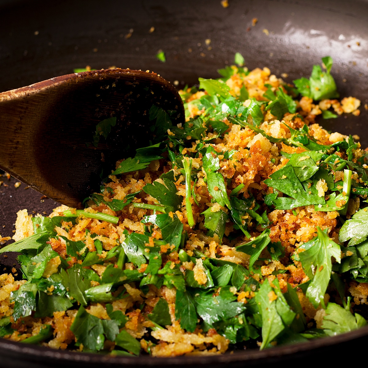 Using a wooden spoon to stir chopped fresh parsley into a skillet with toasted bread crumbs.