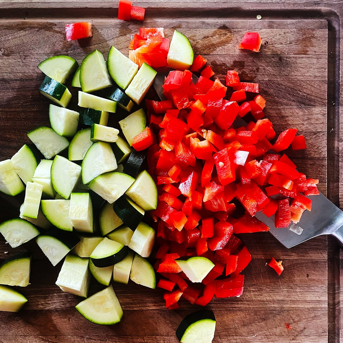 Chopped zucchini and red bell pepper on a wood cutting board.
