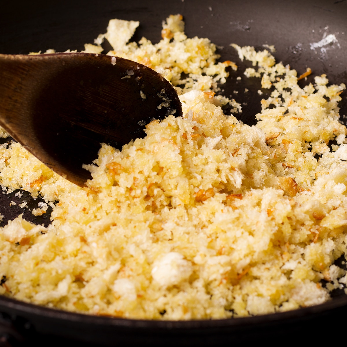 Using a wooden spoon to stir bread crumbs, garlic, and oil in a skillet.