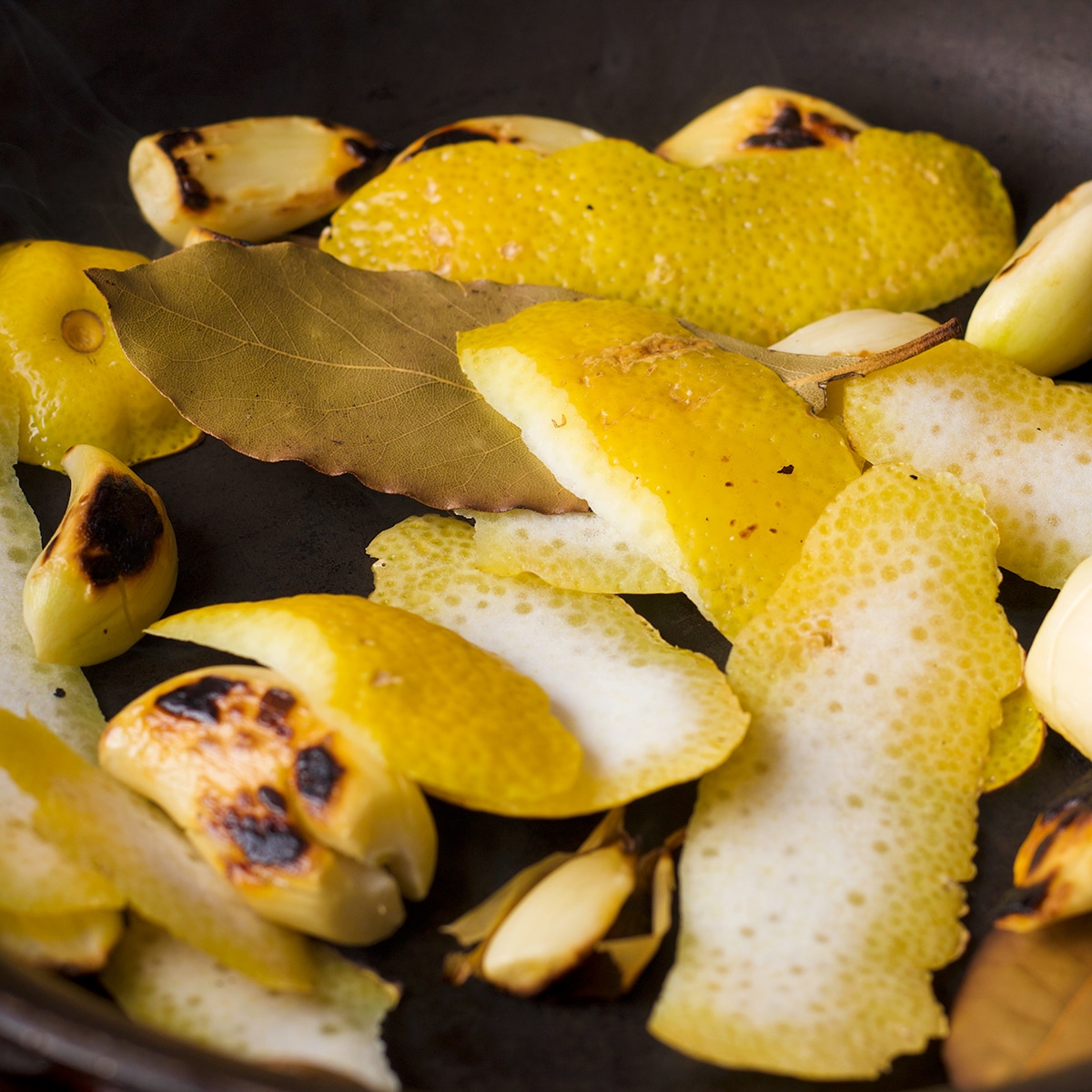 Charred garlic, lemon peel, and bay leaves cooking in a skillet.
