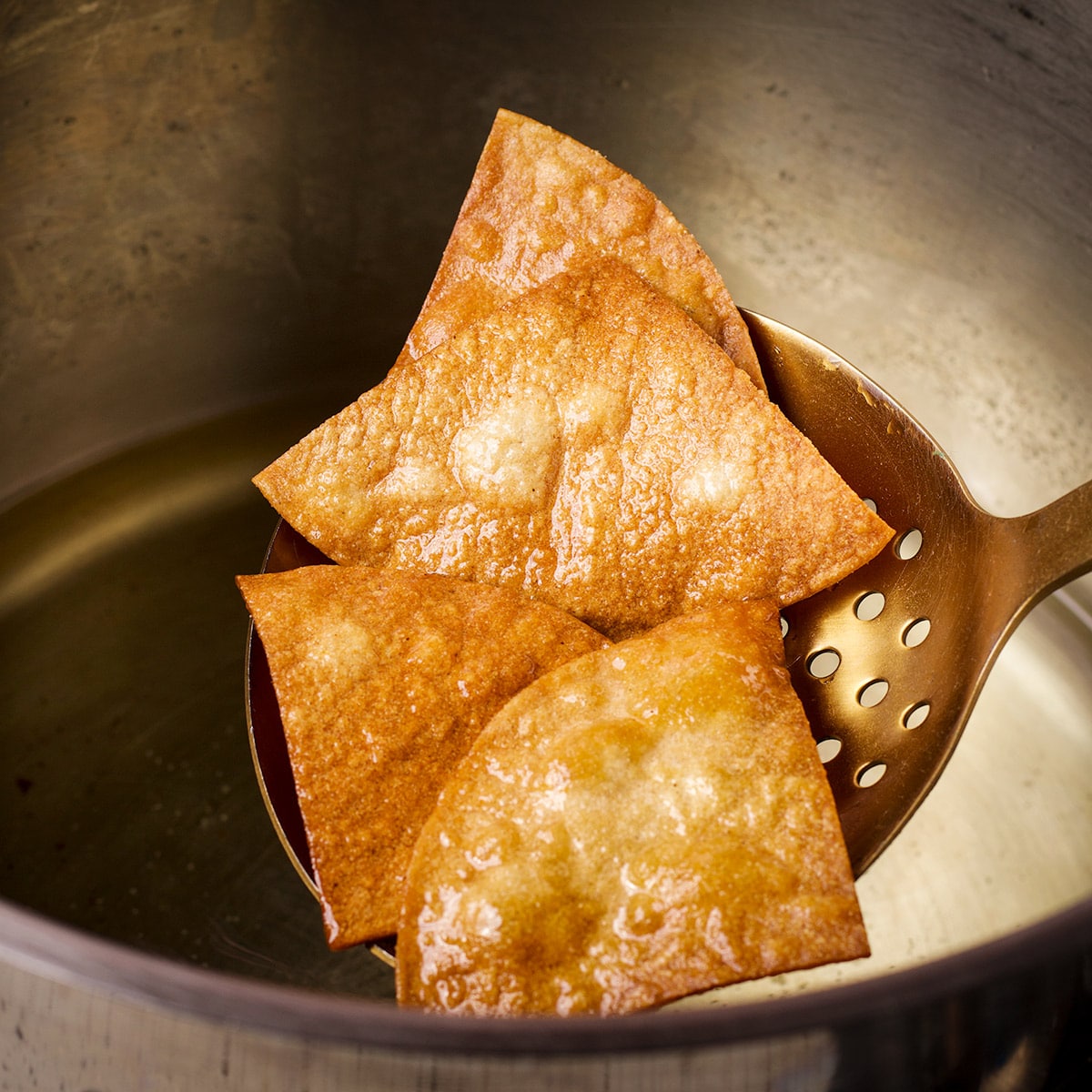 Someone using a large spoon with drain holes to lift corn tortilla chips from a pan of hot oil.