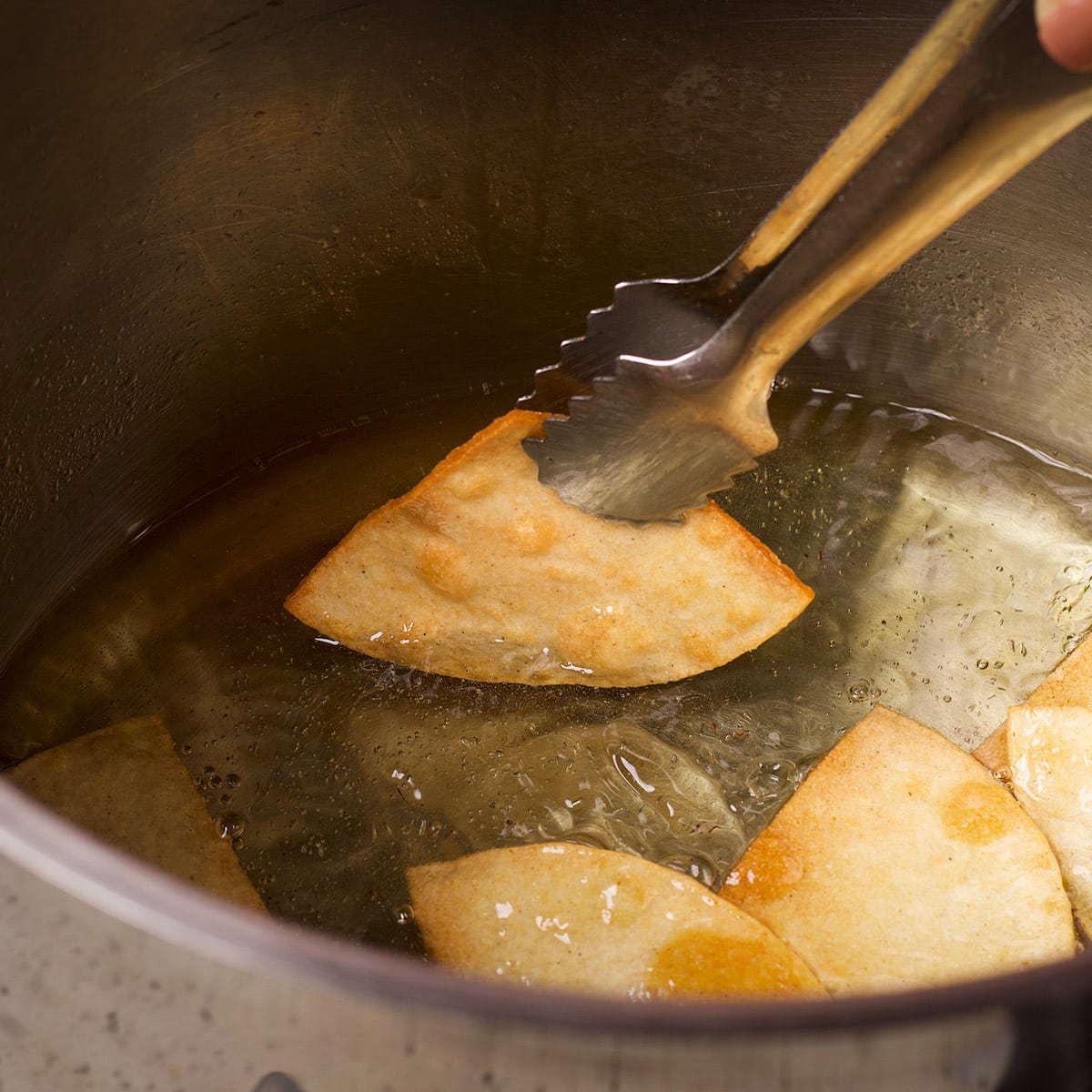 Someone using metal tongs to turn corn tortilla chips over while they cook in hot oil.