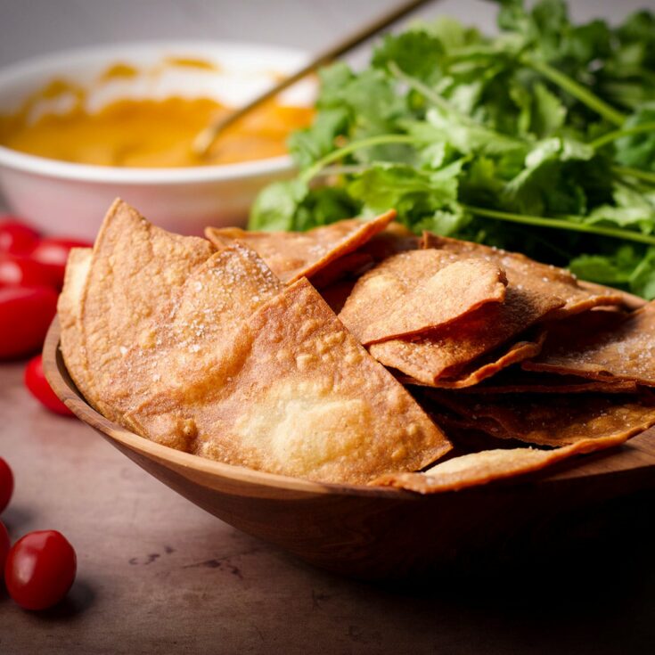 A bowl filled with homemade tortilla chips next to a bowl of nacho cheese sauce, fresh cilantro, and some cherry tomatoes.