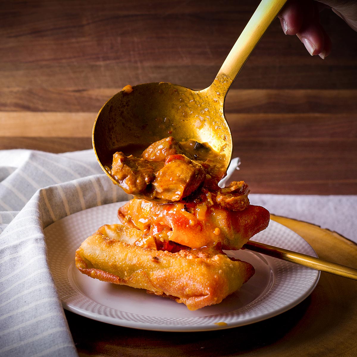 Someone using a gold ladle to smother two chili rellenos with green chili.