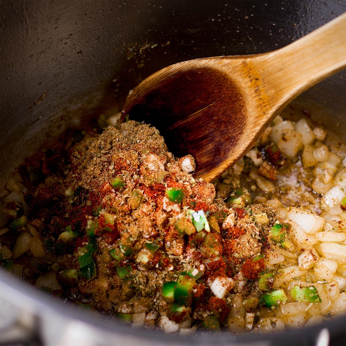 Using a wooden spoon to stir garlic, jalapeño, chili powder, cumin, and oregano into onions cooking in a saucepan.