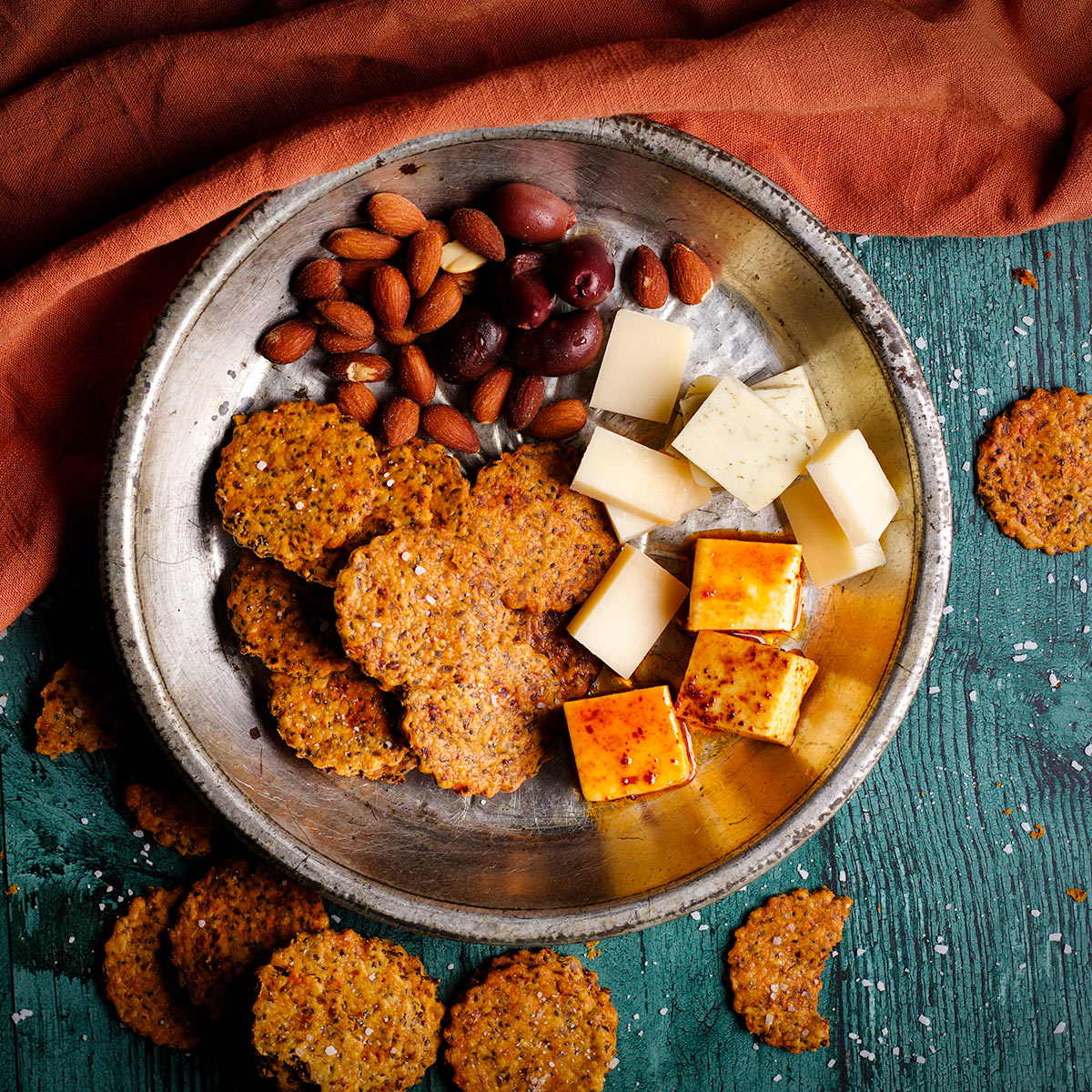 A tin plate filled with homemade crackers, slices of cheese, olives, and almonds.