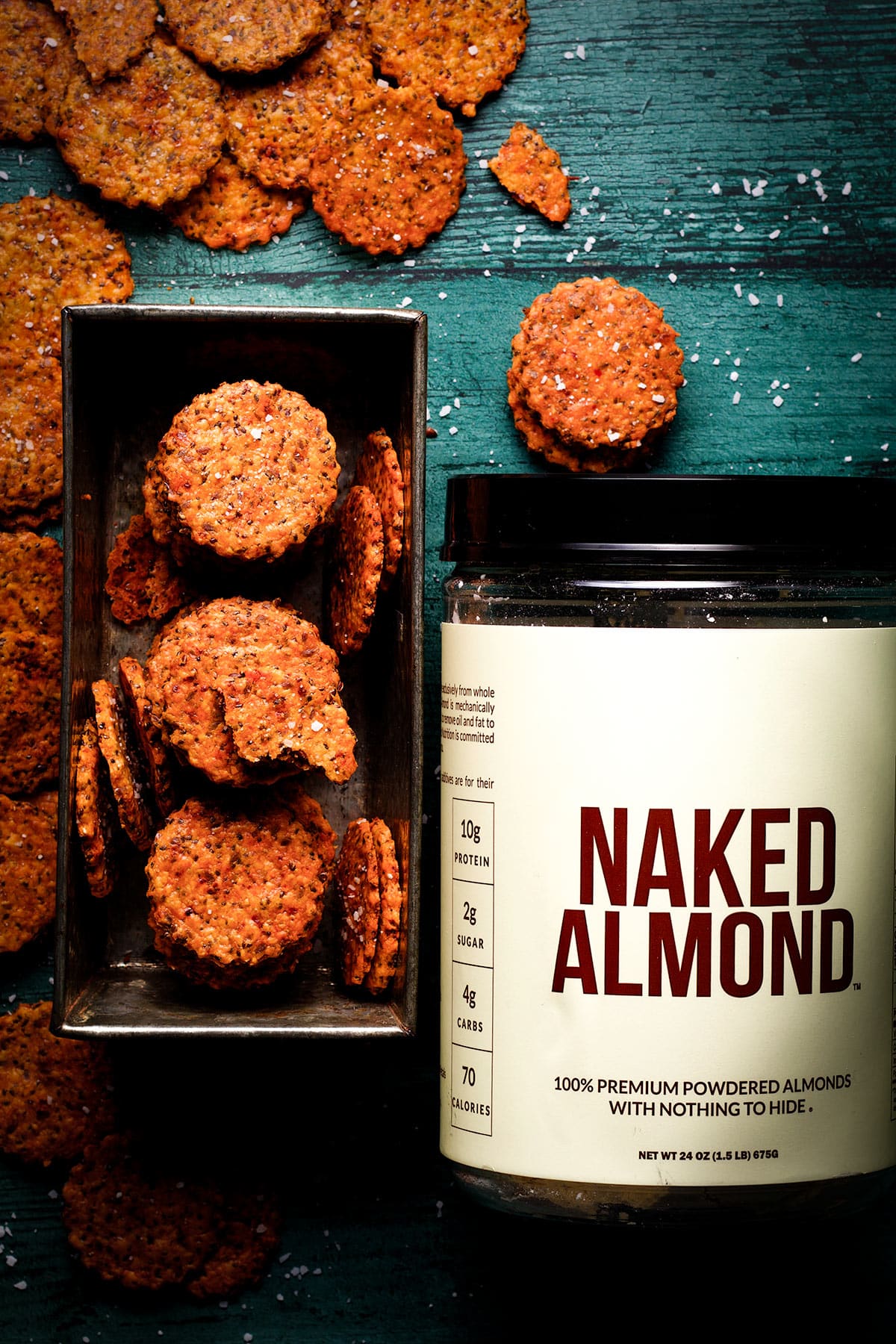 A tin container filled with homemade cheese crackers next to a container of Naked Almond powder.