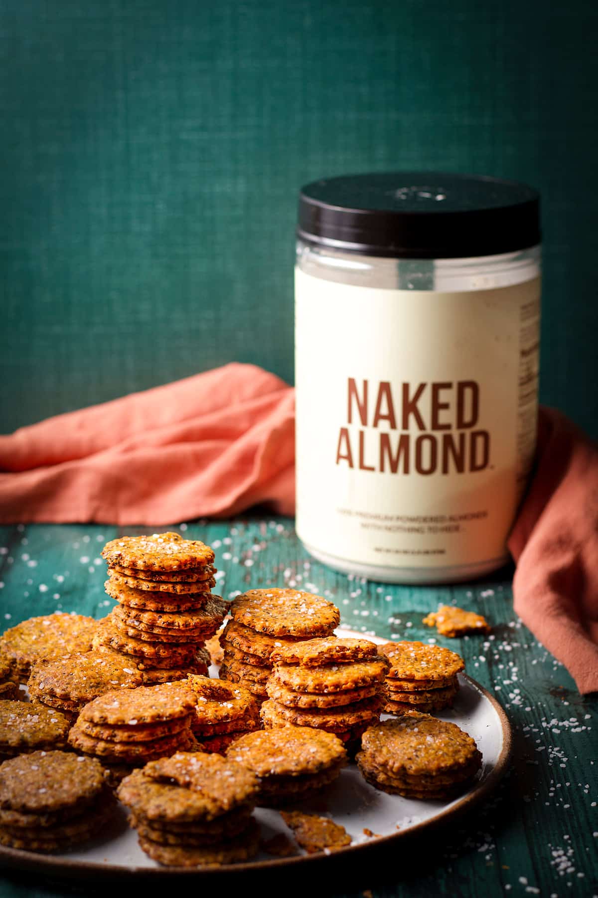Stacks of gluten free almond cheese crackers on a table next to a container of Naked Almond powder.
